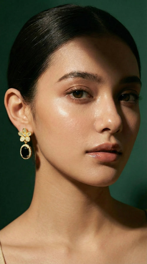 Gold flower-shaped earrings with clear gemstones and dangling oval black gemstone surrounded by small clear stones, worn by a woman.
