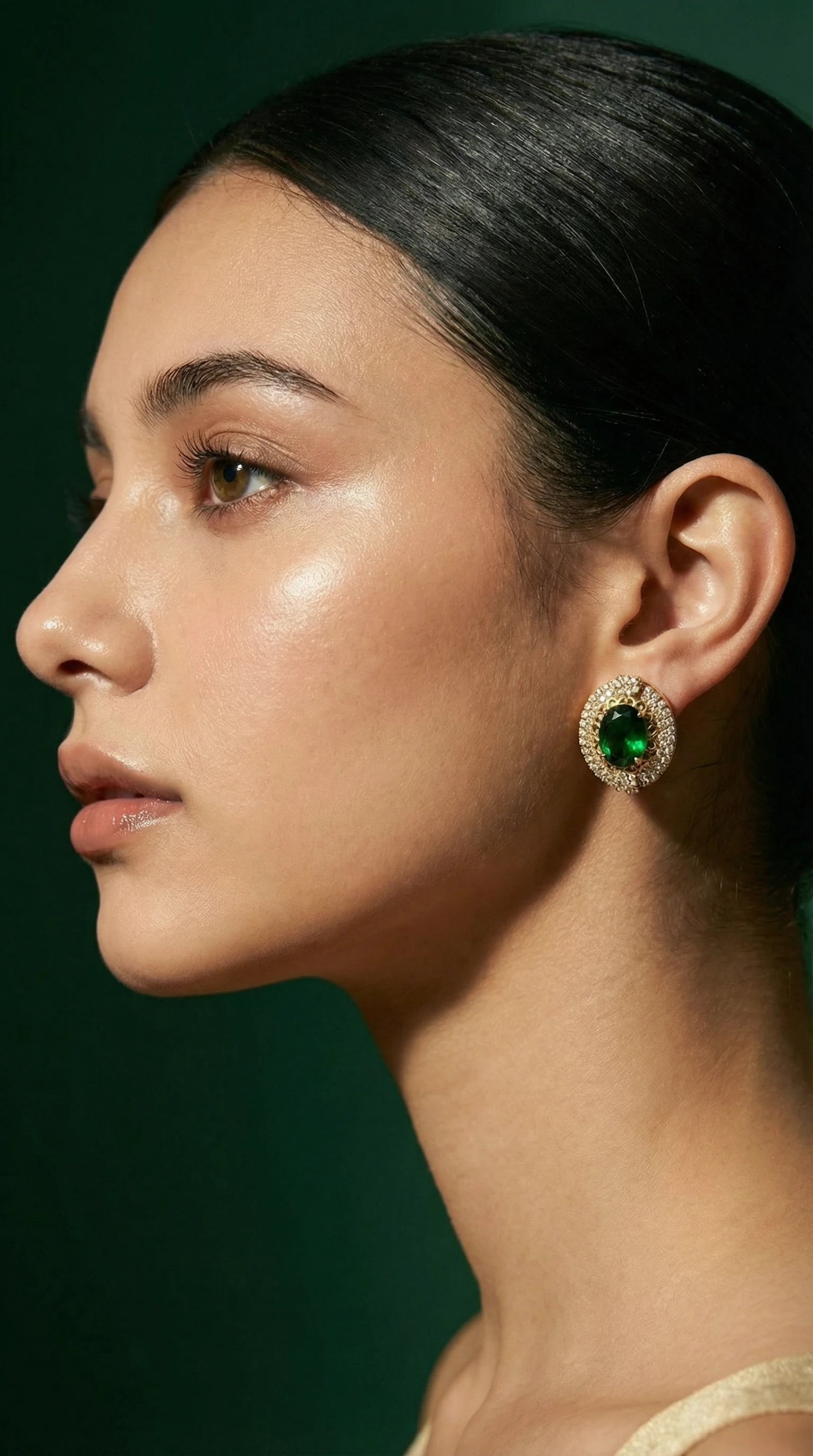 Gold stud earrings with large oval green gemstones surrounded by small clear crystals, worn by a woman.