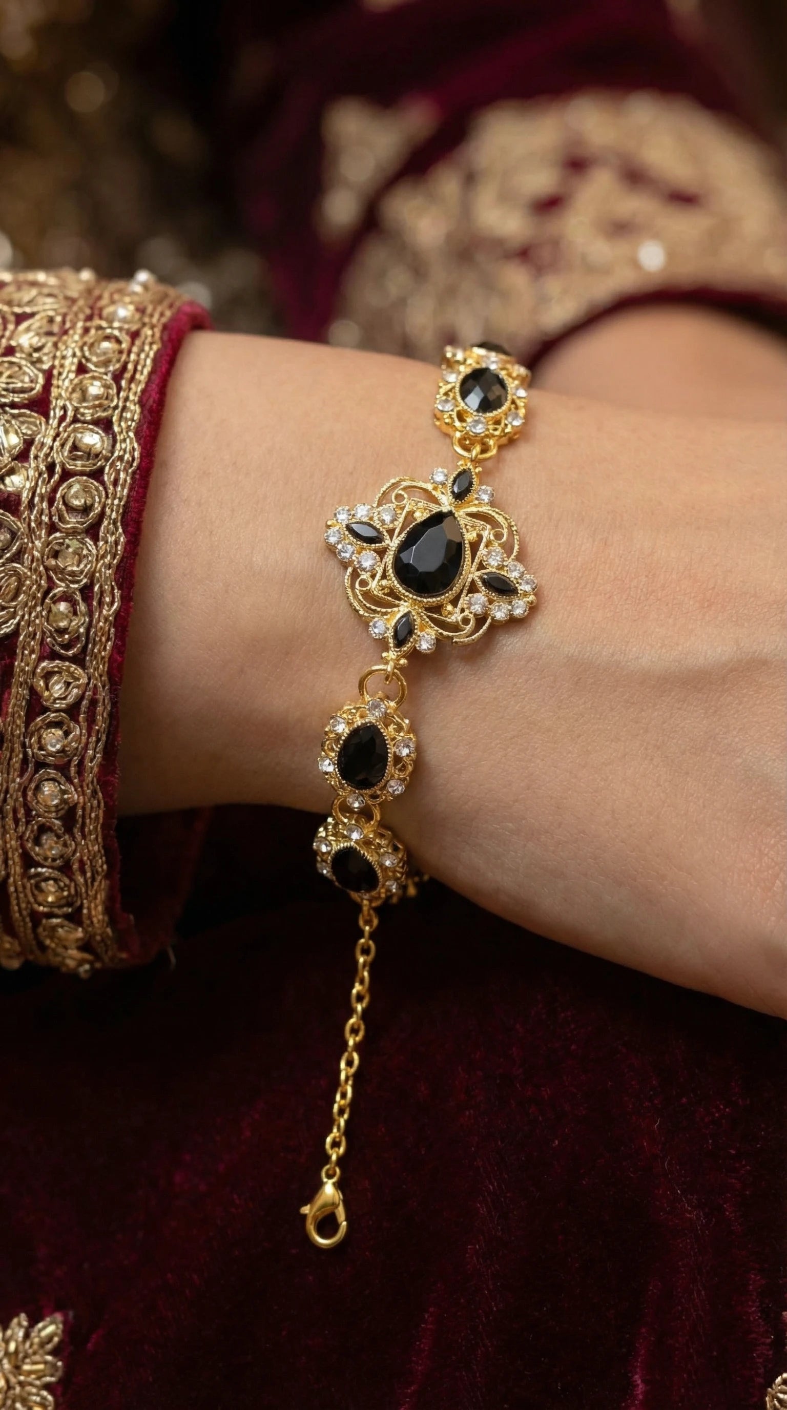 Gold bracelet with intricate design featuring large teardrop and oval black gemstones surrounded by small clear crystals, worn on a wrist with embroidered maroon fabric.