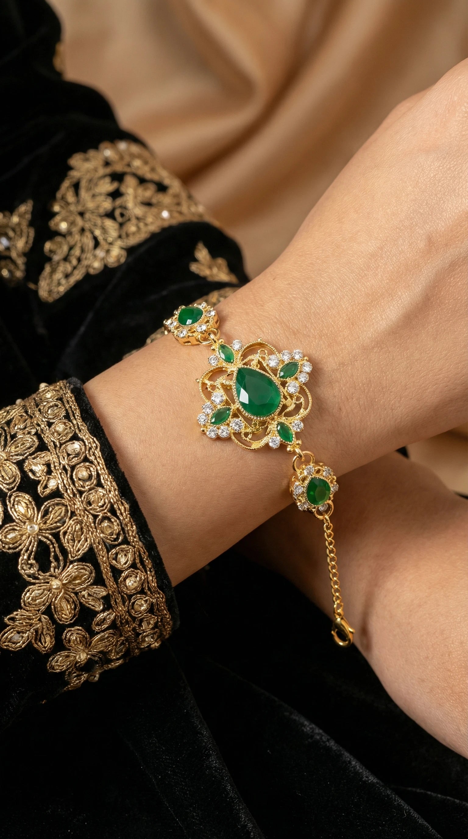 Gold bracelet with intricate design featuring large teardrop and oval green gemstones surrounded by small clear crystals, worn on a wrist with embroidered maroon fabric.