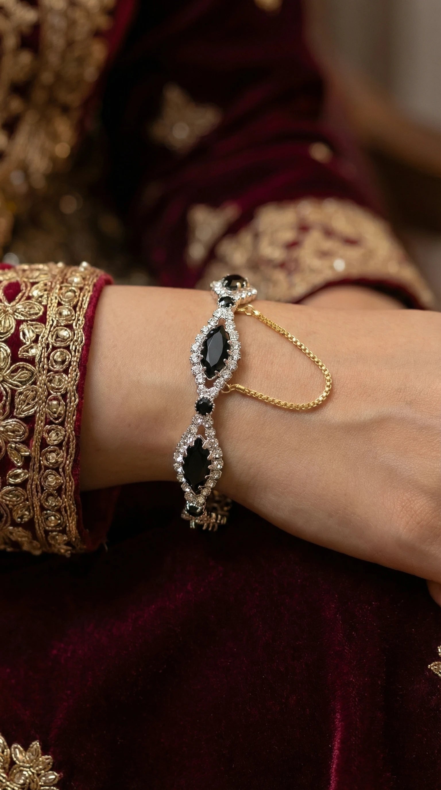 Silver bracelet with marquise-shaped black gemstones surrounded by clear crystals, connected with a gold safety chain, worn on a wrist with embroidered maroon fabric.