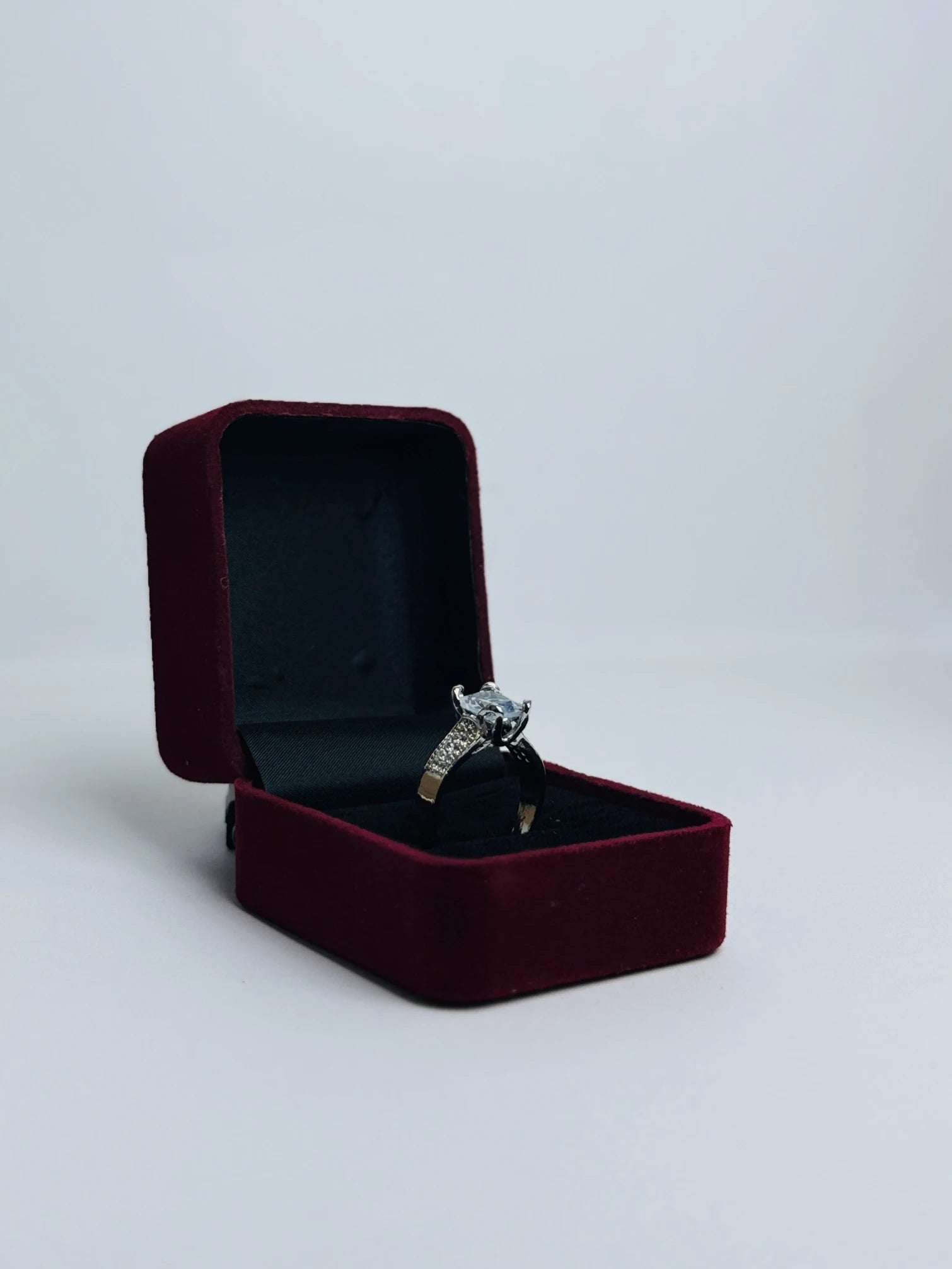 Silver engagement ring with a square-cut clear gemstone and pavé diamond accents on the band, presented in a red velvet ring box.