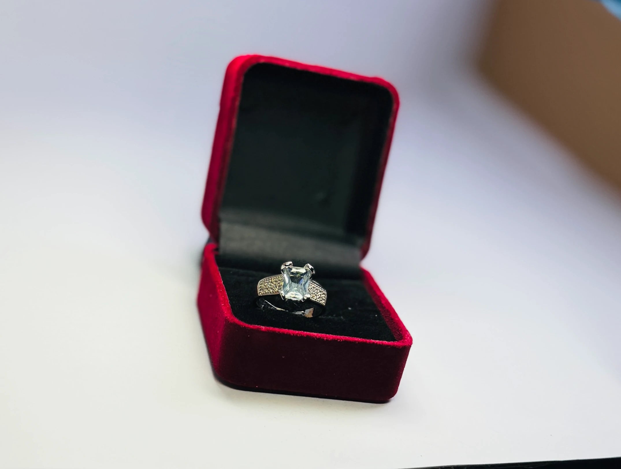 Silver engagement ring with a square-cut clear gemstone and pavé diamond accents on the band, presented in a red velvet ring box.