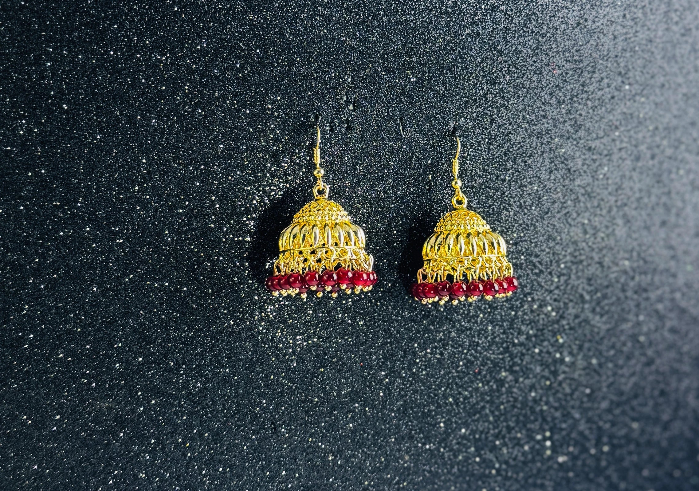 Pair of gold jhumka earrings with intricate dome design and mehroon bead fringes, displayed on a sparkling dark background.