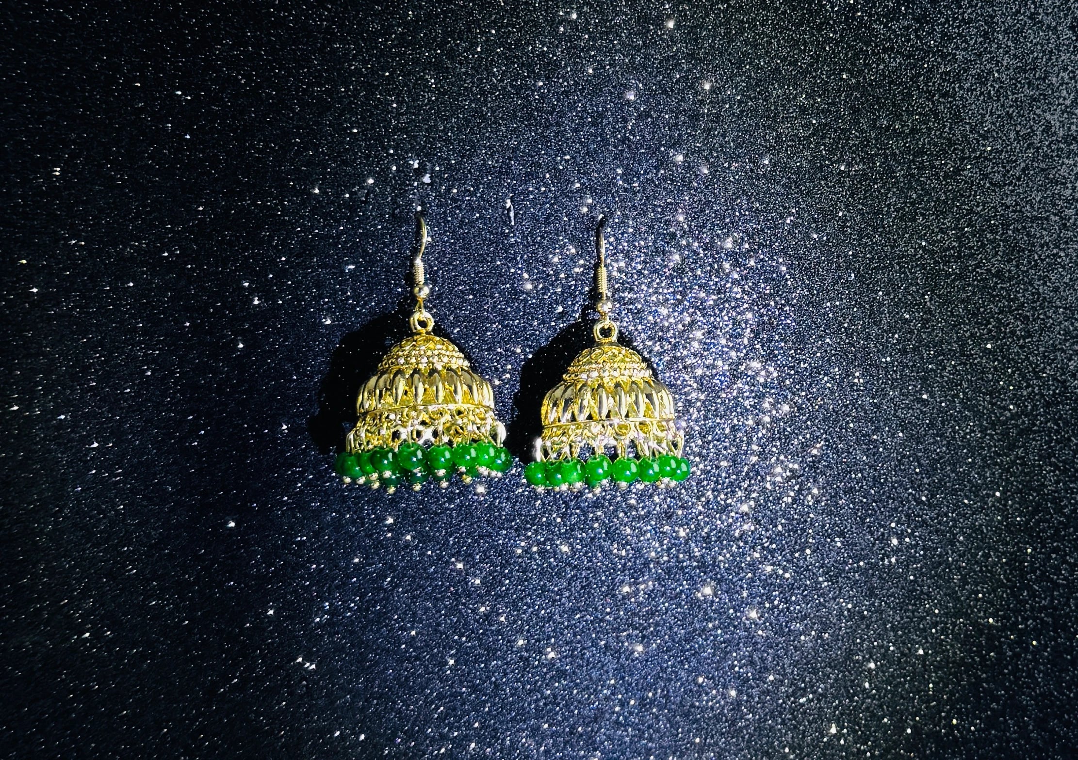 Pair of gold jhumka earrings with intricate dome design and green bead fringes, displayed on a sparkling dark background.