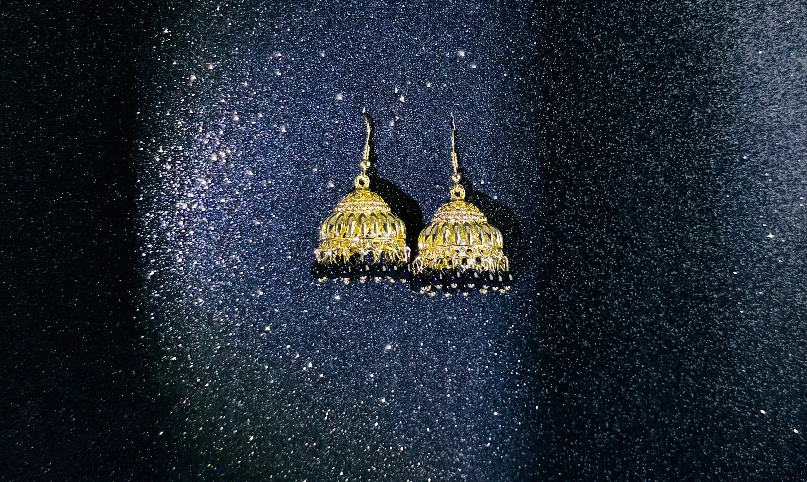 Pair of gold jhumka earrings with intricate dome design and black bead fringes, displayed on a sparkling dark background.