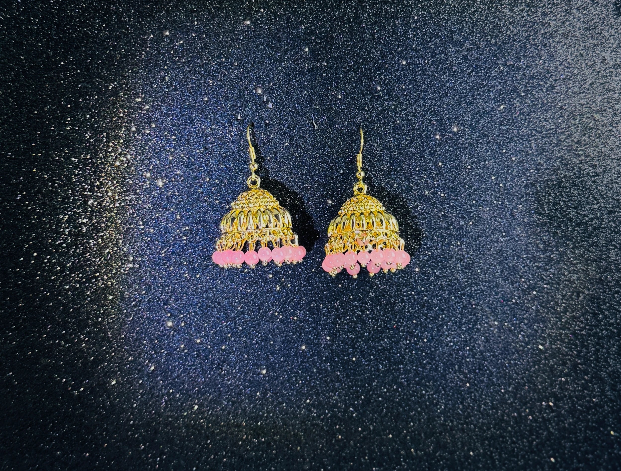 Pair of gold jhumka earrings with intricate dome design and pink bead fringes, displayed on a sparkling dark background.