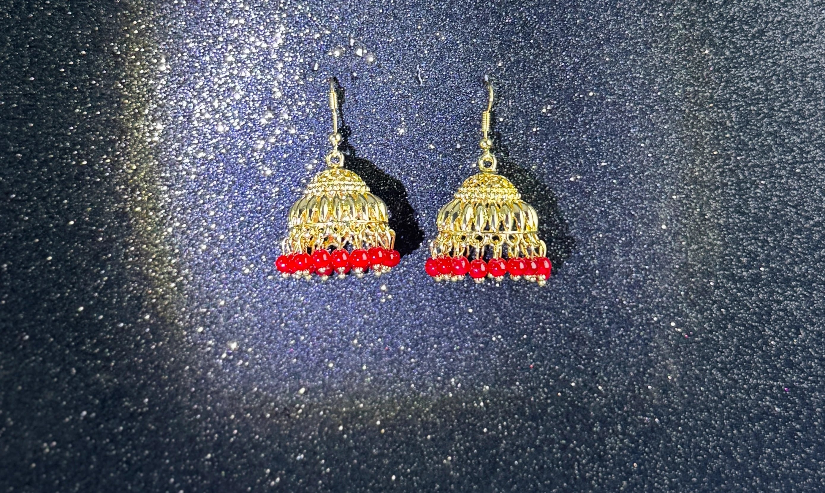 Pair of gold jhumka earrings with intricate dome design and red bead fringes, displayed on a sparkling dark background.