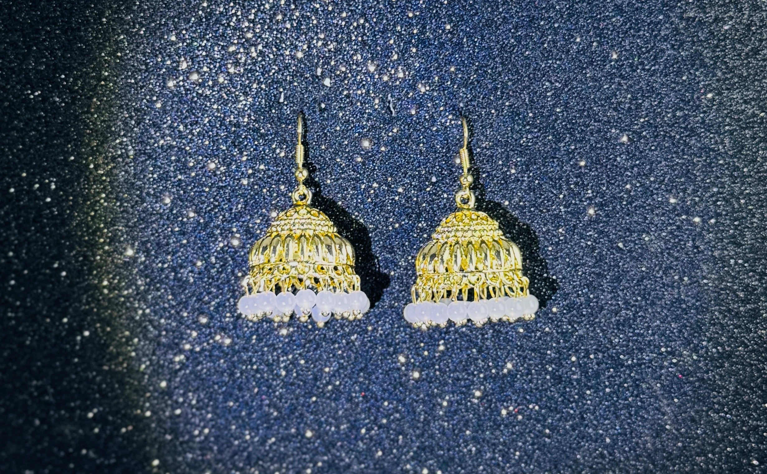 Pair of gold jhumka earrings with intricate dome design and white bead fringes, displayed on a sparkling dark background.