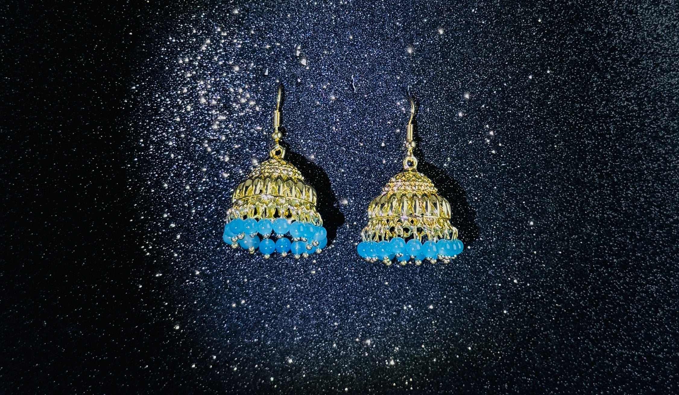 Pair of gold jhumka earrings with intricate dome design and blue bead fringes, displayed on a sparkling dark background.