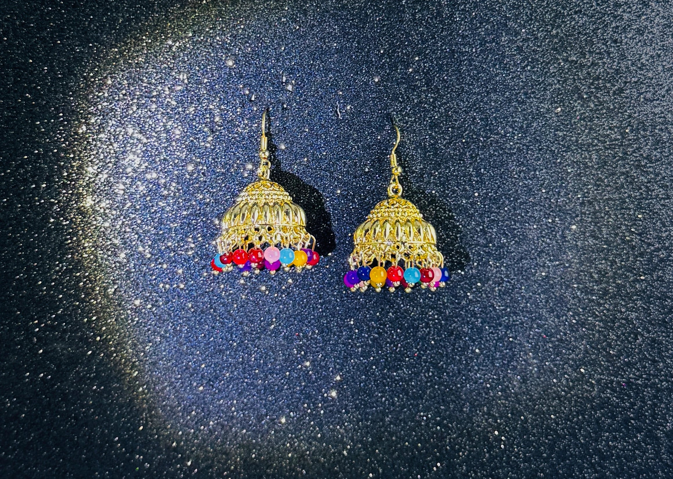 Pair of gold jhumka earrings with intricate dome design and multicolor bead fringes, displayed on a sparkling dark background.