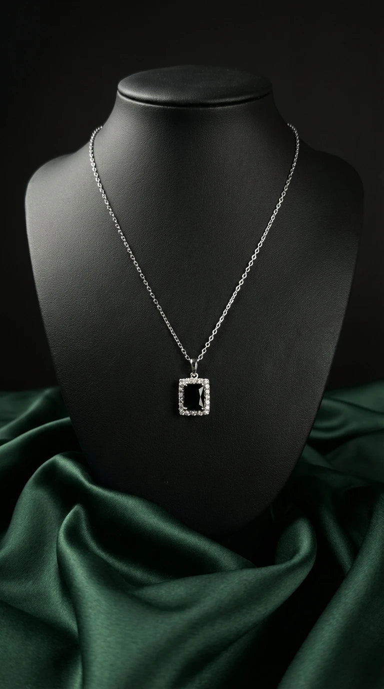 Silver necklace with a rectangular black gemstone pendant surrounded by small clear crystals, displayed on a black bust with dark green fabric background.