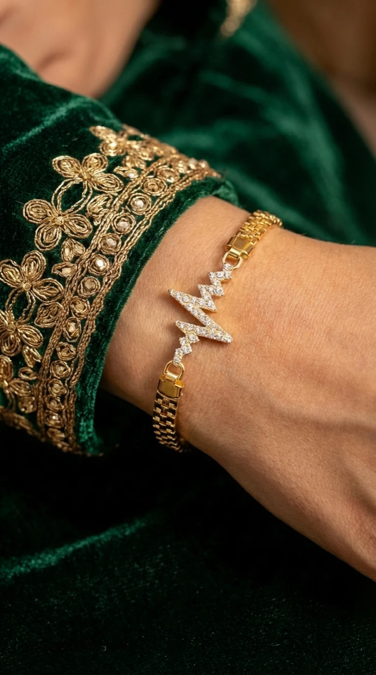 Gold bracelet with a zigzag lightning bolt design encrusted with clear gemstones, worn on a wrist with green embroidered fabric.